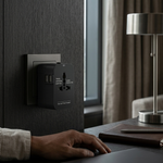 A black universal travel power adapter plugged into a wall socket on a dark wood desk, with a hand resting nearby and a laptop in the foreground.