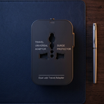 A black dual USB travel adapter with a plug-shaped cutout, placed on a dark navy desk mat with a silver pen beside it on a dark wood background.