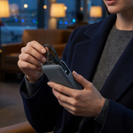 A woman in a navy blazer and gray sweater sits in a dimly lit lounge area, holding a smartphone and attaching a black cable to its port.