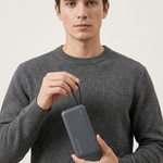 A man wearing a dark gray knit sweater holds a dark gray rectangular power bank with a carrying strap against a light gray studio background.