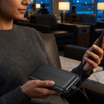 A woman in a gray sweater sits in a modern lounge setting, holding a black rectangular electronic device and connecting a smartphone charger to it.