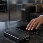 A black rectangular portable power bank sits on a dark marble table next to a smartphone, with a person's hand reaching toward the phone. The background shows a modern office lounge setting with a sofa and glass partitions.
