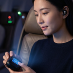 A woman seated in a plane cabin holds a small black electronic device with a blue display, wearing a matching black earbud. The scene is dimly lit with cabin lights in the background.