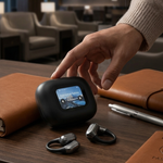 A person's hand reaches toward a black wireless earbud charging case on a wooden desk, with two wireless earbuds placed below it. The scene includes brown leather notebooks, a pen, and a softly blurred lounge interior in the background.