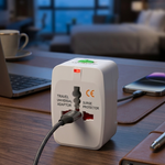 A white travel universal adapter with a built-in surge protector and a braided black power cord, shown plugged into a wooden desk in a blurred modern office or hotel room setting.