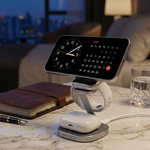 A modern wireless charging stand holding a smartphone and a pair of white earbuds, placed on a marble tabletop in a softly lit bedroom setting.