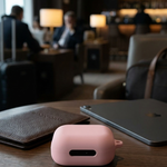 A small pink rectangular accessory case sits on a wooden table in a blurred cafe or lounge setting, with a brown leather wallet and a closed silver laptop nearby.