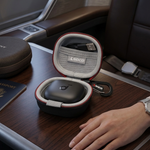 A close-up of a person's hand resting on a premium airplane tray table next to an open MagSafe-compatible protective case for wireless earbuds, with a matching round case on the tray table.