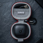 An open round protective earbud charging case with red trim and gray interior, shown on a dark textured surface. A black charging case lid sits inside, with a USB-C cable in the upper compartment and a small branded keychain tag.