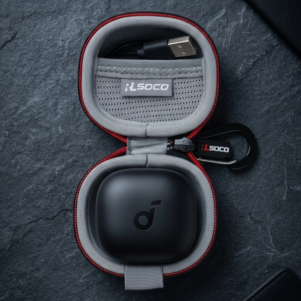 An open round protective earbud charging case with red trim and gray interior, shown on a dark textured surface. A black charging case lid sits inside, with a USB-C cable in the upper compartment and a small branded keychain tag.