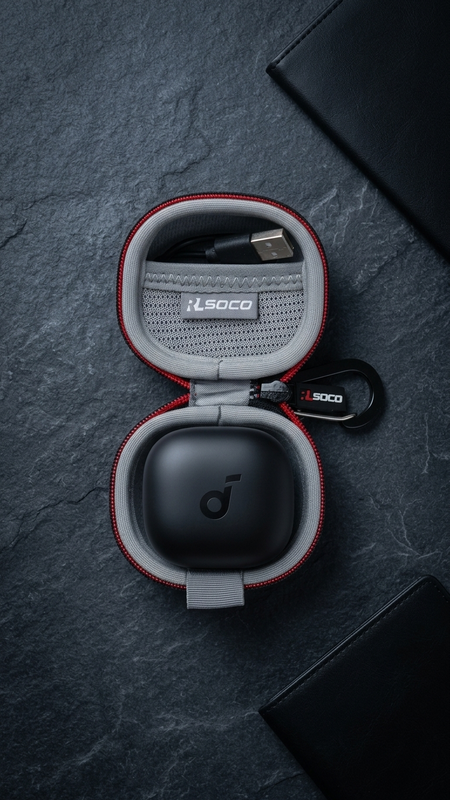 An open round protective earbud charging case with red trim and gray interior, shown on a dark textured surface. A black charging case lid sits inside, with a USB-C cable in the upper compartment and a small branded keychain tag.