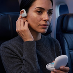 A woman in a gray sweater sits in an airplane seat, inserting a white earbud into her ear while holding an open white charging case.