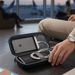 A black zippered travel organizer case is open on a dark leather surface, holding a white power bank and white cable. A person's hand rests on the case, with an airport lounge setting, airplane windows, and seated passengers blurred in the background.