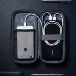 A black zippered portable charging case opened flat on a dark textured surface, containing a white power bank on the left and a black power bank on the right with white cables. The case is styled with pens and a leather notebook around it.