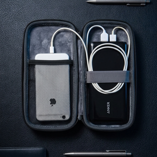 A black zippered portable charging case opened flat on a dark textured surface, containing a white power bank on the left and a black power bank on the right with white cables. The case is styled with pens and a leather notebook around it.