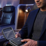 A man in a navy blazer and gray sweater sits on an airplane, holding an open gray carrying case with a black rectangular device inside. The airplane cabin is visible in the background with blue lighting.