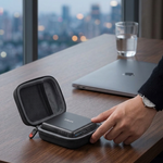 A person's hand is shown placing a small black portable device into an open gray zippered carrying case on a wooden desk. In the background, a closed silver laptop and a glass of water sit near a large window with a blurred city skyline outside.