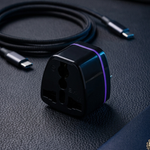 A black universal travel adapter with a purple accent band, shown next to a coiled black USB-C cable, a silver stylus, and a blue passport on a dark textured surface.