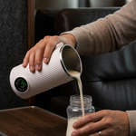 A person pours milk from a white ribbed electric milk pitcher with a digital display, into a clear baby bottle, set on a wooden table in a modern interior.