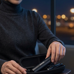 A woman in a dark gray turtleneck sweater opens a black zippered carrying case on a wooden table, revealing a sleek black hair styling tool inside, with a softly blurred night city window in the background.