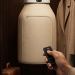 A beige hanging garment storage bag with a white top cap is shown inside a wooden closet, with a hand holding a black remote control nearby and a tan coat hanging on the right.