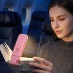 A woman in an airplane seat reads a book with a pink portable clip-on light illuminating the pages.