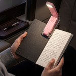 A person seated in a train or airplane reading a book, with a small pink portable book light clipped onto the open book. The scene is lit by the book light, showing the illuminated reading area against dark upholstery and a window in the background.