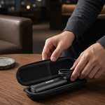 A person's hands open a black zippered carrying case on a wooden table, revealing a sleek black hair styling tool with a coiled cord inside. The background shows a softly blurred modern indoor setting with warm lighting.