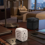A white universal travel power adapter with multiple international outlets sits on a dark wooden desk in a modern hotel room. The desk is styled with a silver desk lamp, a black zippered case, a tablet, and notebooks, with a bed and city view visible in the background.