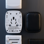 A white multi-port travel adapter with multiple USB-C and USB-A ports, shown alongside its reverse side, a black zippered carrying case, and a black leather wallet on a dark textured background.