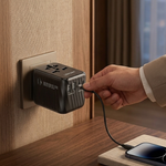 A black universal travel adapter plug is shown being plugged into a wall socket on a wooden desk, with a hand in a beige suit sleeve holding the connector. A smartphone is charging on a wooden stand nearby.