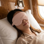 A woman lying down on a bed or lounge chair wearing a light pink sleep mask and beige pajamas, with pillows, wood-paneled walls, and a window visible.