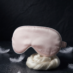 A pale blush pink satin sleep mask with white piping and a small embroidered logo, displayed on a white decorative stand against a dark textured background with scattered feathers and lavender sprigs.