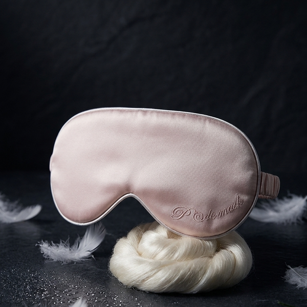 A pale blush pink satin sleep mask with white piping and a small embroidered logo, displayed on a white decorative stand against a dark textured background with scattered feathers and lavender sprigs.