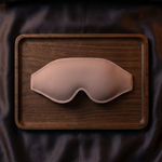 A pale blush pink molded sleep mask resting on a rectangular dark brown wooden tray, which is placed on dark satin bedding.