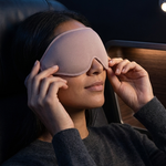 A woman wearing a light pink sleep mask rests in a dark airplane seat, adjusting the mask with both hands.