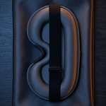 A dark gray padded travel neck pillow with a black strap, presented on a dark textured fabric background.