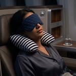 A woman reclining in an airplane seat with her eyes closed, wearing a dark blue quilted sleep mask and a black-and-white striped neck pillow.