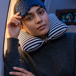 A woman wearing a dark blue quilted travel eye mask and a black-and-white striped neck pillow, resting her head on an airplane seat.