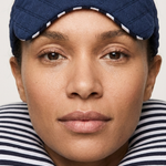 A close-up of a woman wearing a navy blue quilted travel eye mask and a matching navy and white striped neck pillow against a light gray background.