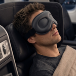 A man resting in an airplane seat with a dark gray sleep mask on his eyes, wrapped in a beige blanket.