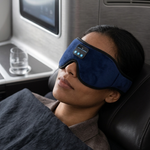A person reclining in a seat, wearing a dark blue sleep mask with small illuminated control buttons. The setting appears to be a travel or lounge chair with an in-seat screen and tray table in the background.