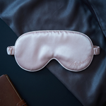 A light pink satin sleep mask with elastic straps, featuring a subtle white embroidered logo, displayed on a dark navy blue silk sheet next to a brown leather notebook.