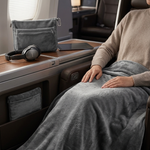 A person reclining in an airplane seat, wrapped in a plush gray blanket. A matching gray pouch and headphones rest on the tray table beside them.