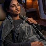 A woman reclining in a luxurious airplane or train seat, wrapped in a plush gray blanket over a dark gray top.