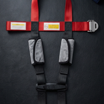 A red and black safety harness laid flat on a dark textured surface, with gray padded shoulder straps and a silver metal buckle. A folded dark blue cloth is partially visible in the top right corner.