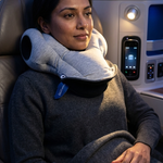 A woman seated in an airplane cabin wearing a light gray travel neck pillow around her neck, with a dark gray sweater and blanket covering her lap.