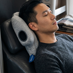 A man rests his head on a gray contoured travel neck pillow inside an airplane cabin next to a window.