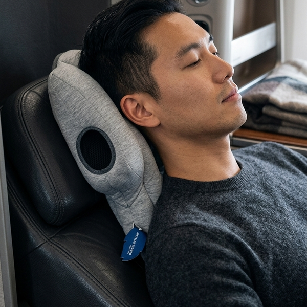 A man rests his head on a gray contoured travel neck pillow inside an airplane cabin next to a window.