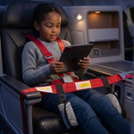 A young girl sits in a reclined airplane seat, fastened with a red safety harness, holding a tablet device. The cabin interior is dimly lit with blue ambient lighting.